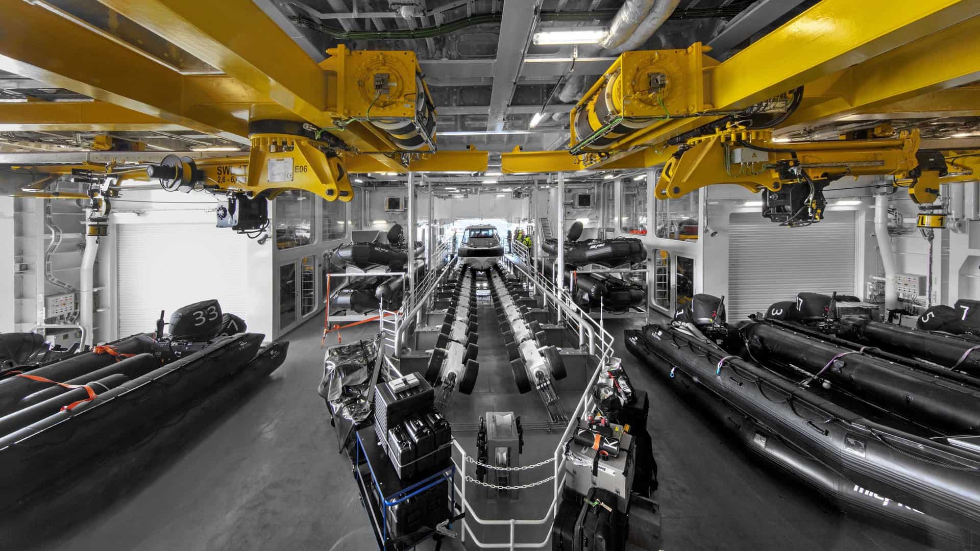 An internal marina hangar aboard an expedition ship features yellow overhead cranes, rows of black inflatable Zodiac boats, and a central roller launch ramp for tender craft.