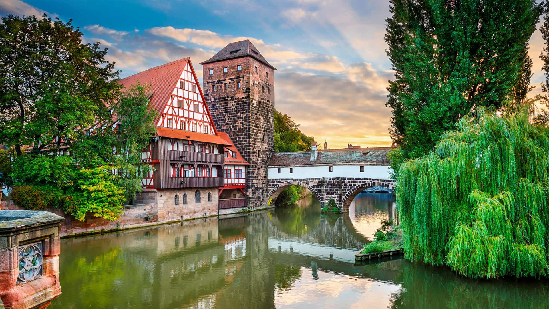 A historic waterfront view featuring a half-timbered building, a tall stone watchtower, and a stone arch bridge spanning a calm canal, flanked by lush green trees.