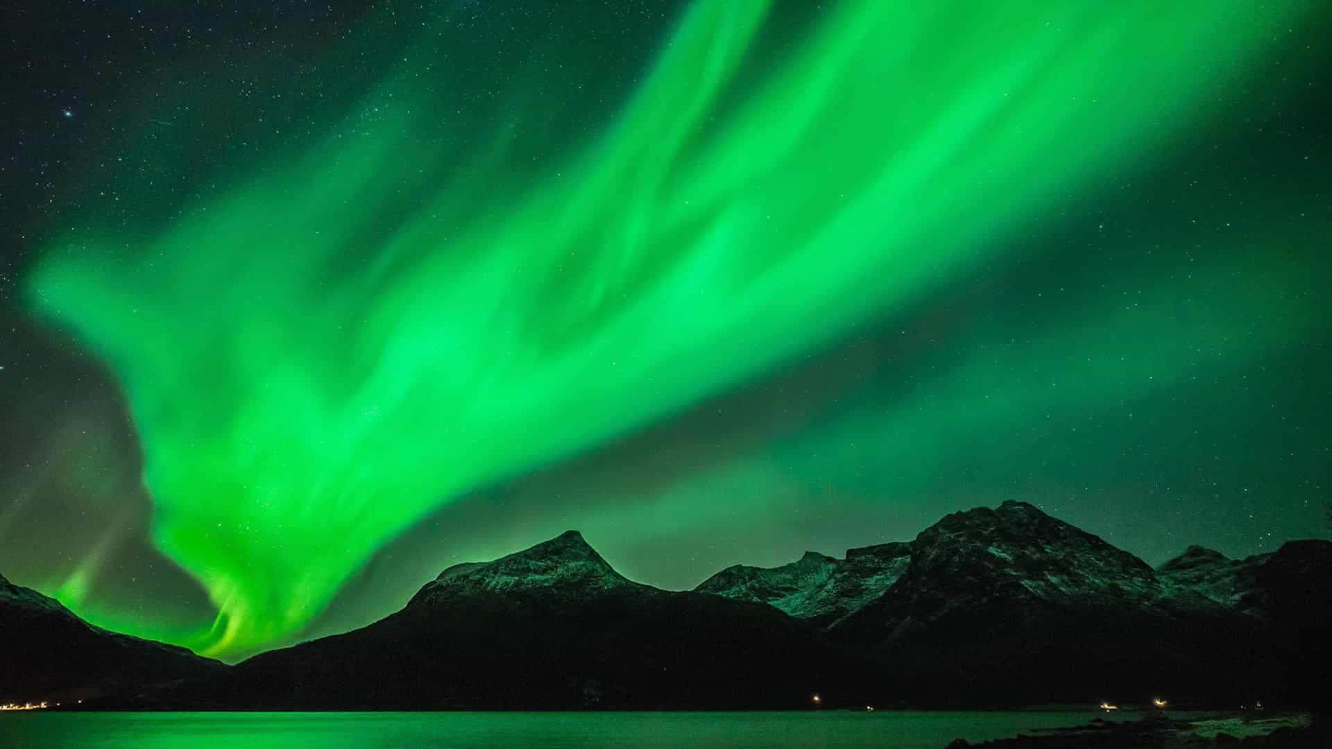 The stunning Northern Lights, or Aurora Borealis, visible over a calm body of water and snow-capped mountains in Tromsø, Norway, a highlight of Viking Ocean cruises to the Arctic Circle.