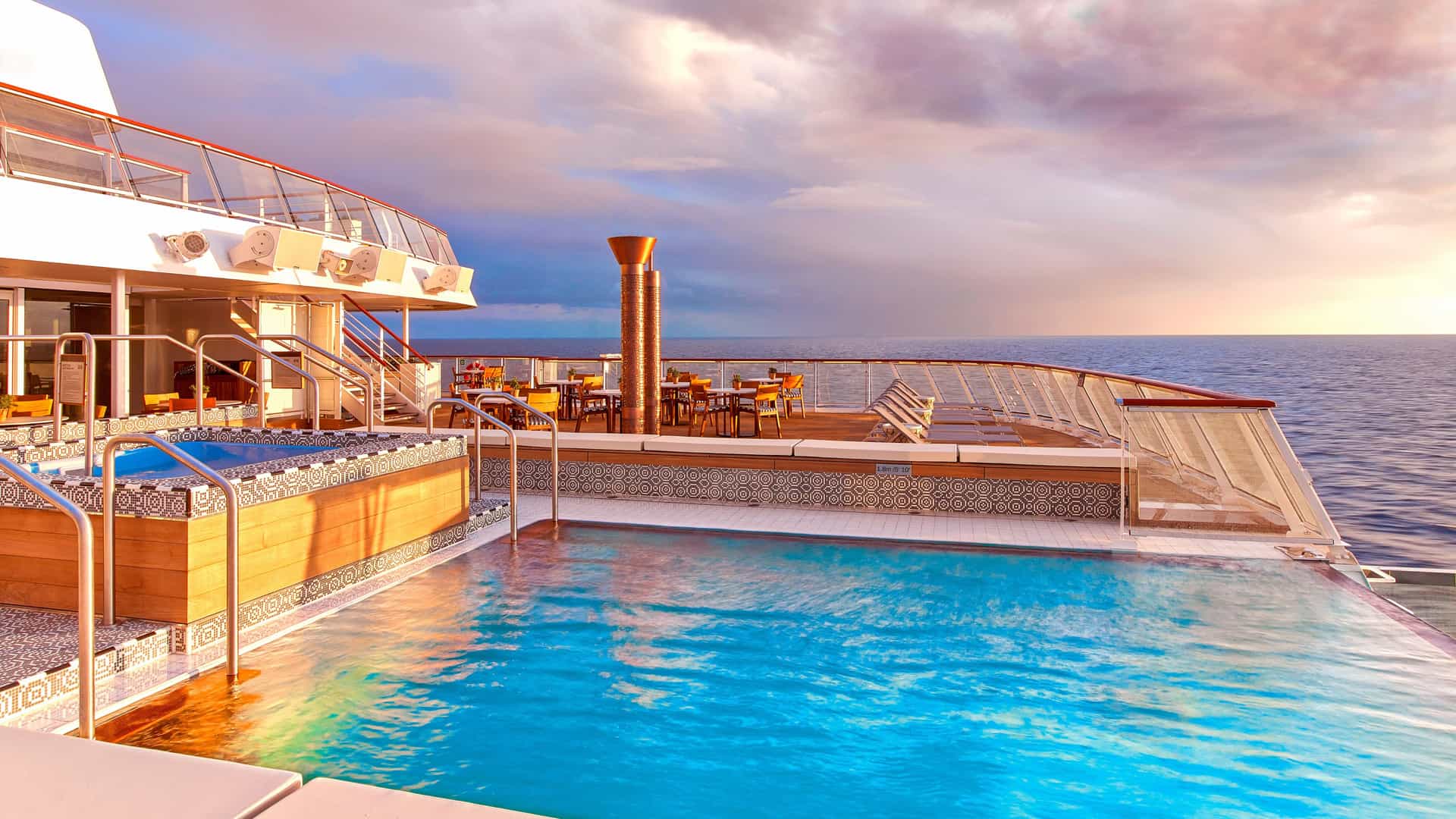 An infinity pool and hot tub on the wooden deck of a cruise ship at sunset. The glowing blue water of the pool appears to merge with the calm ocean on the horizon under a dramatic sky of pink and orange clouds.