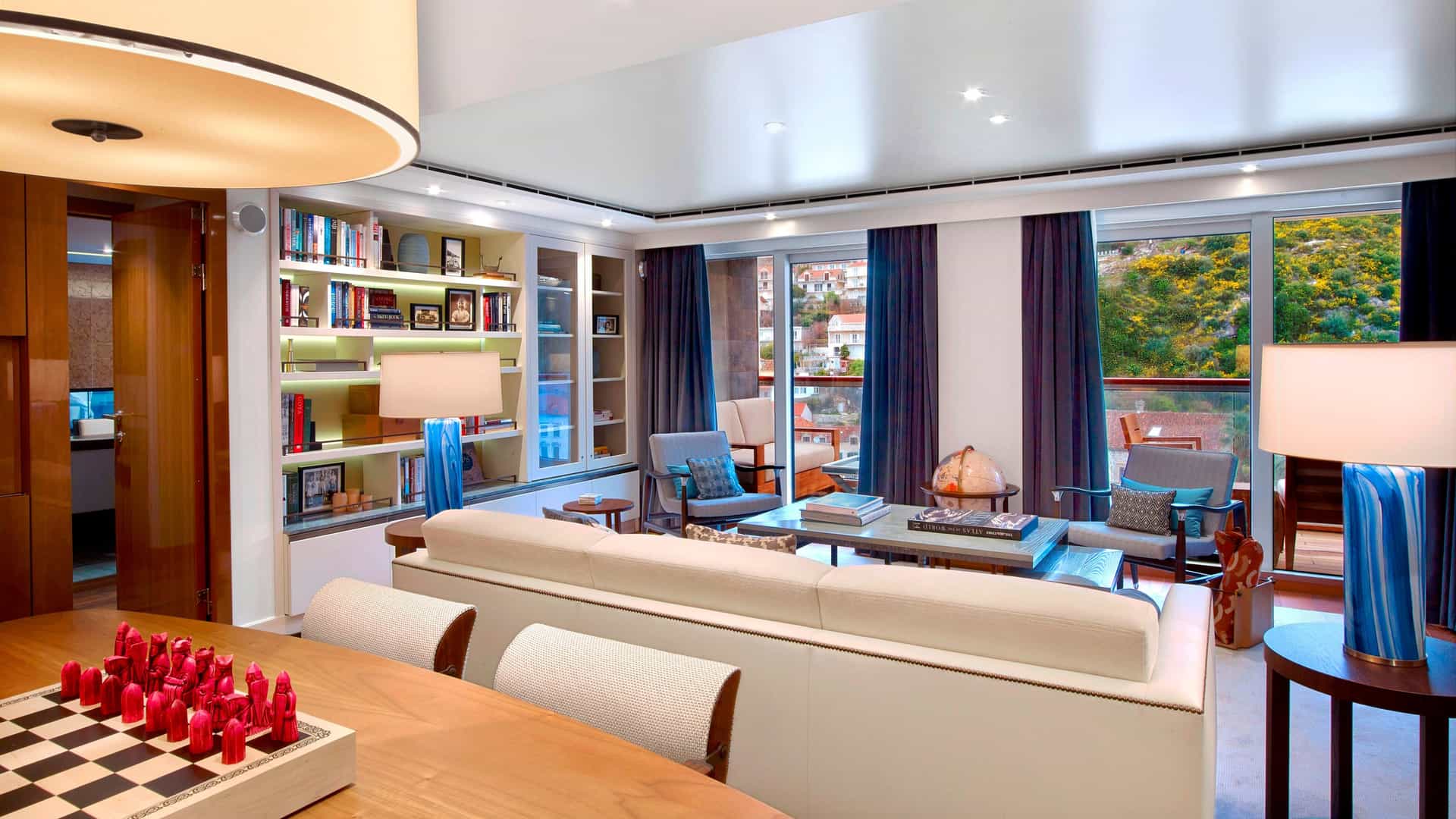 A wide shot of the sitting area of the Owner's Suite aboard Viking Ocean Cruises. There's a chessboard with red pieces on top of a table. A white couch and living area behind with huge windows that let in sun light.
