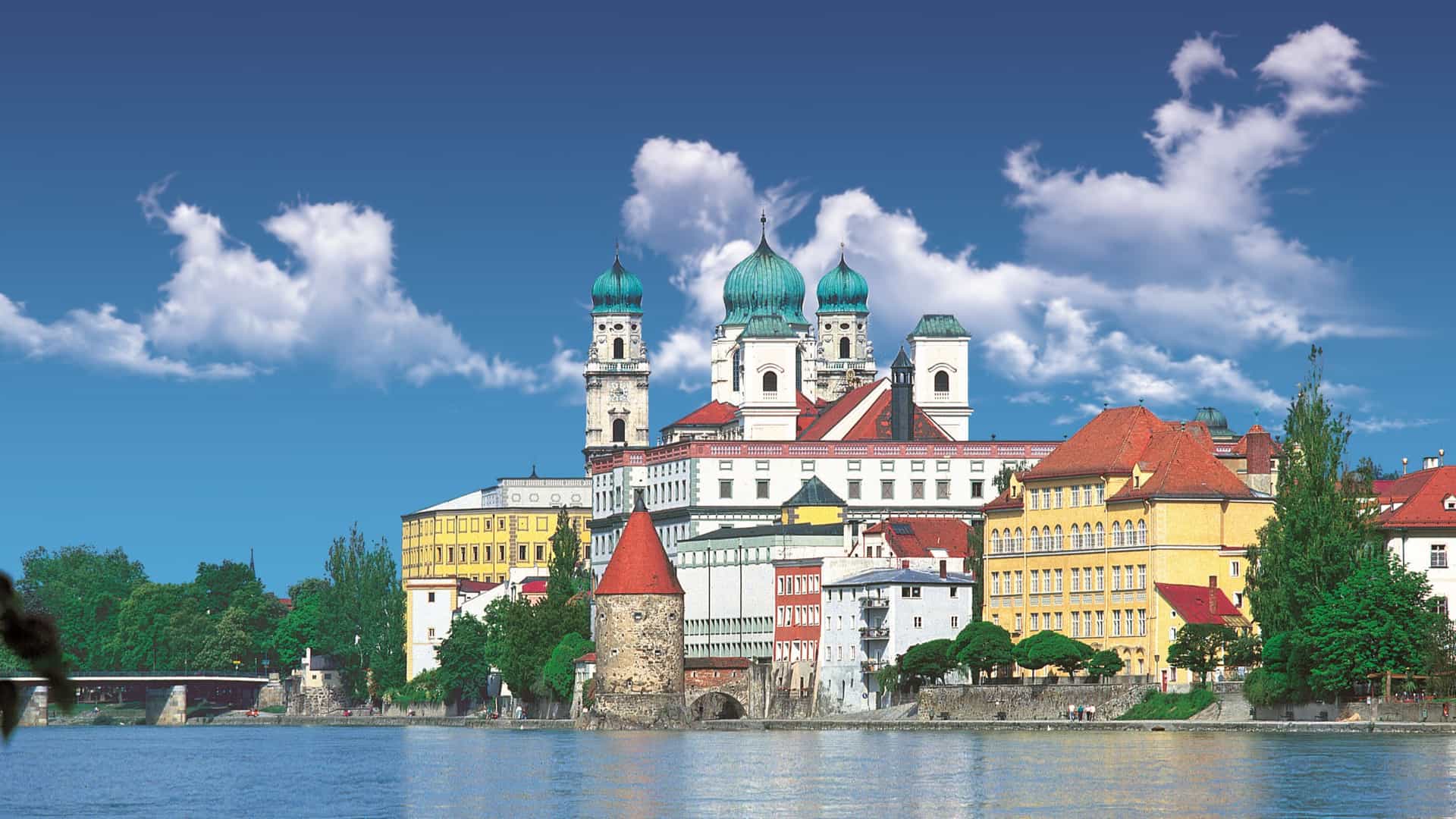 A scenic view of Passau featuring a grand cathedral with teal domes situated along a riverbank under a bright blue sky with scattered clouds.