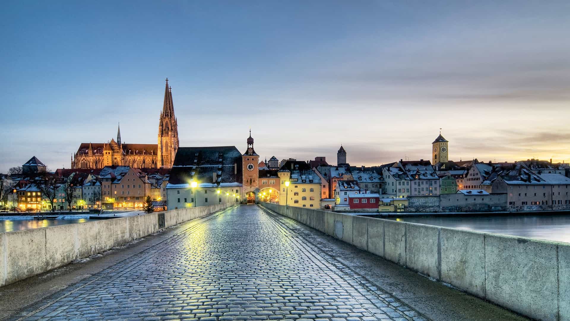 A wet cobblestone stone bridge leading toward the historic town skyline and cathedral spire of Regensburg under a twilight sky.