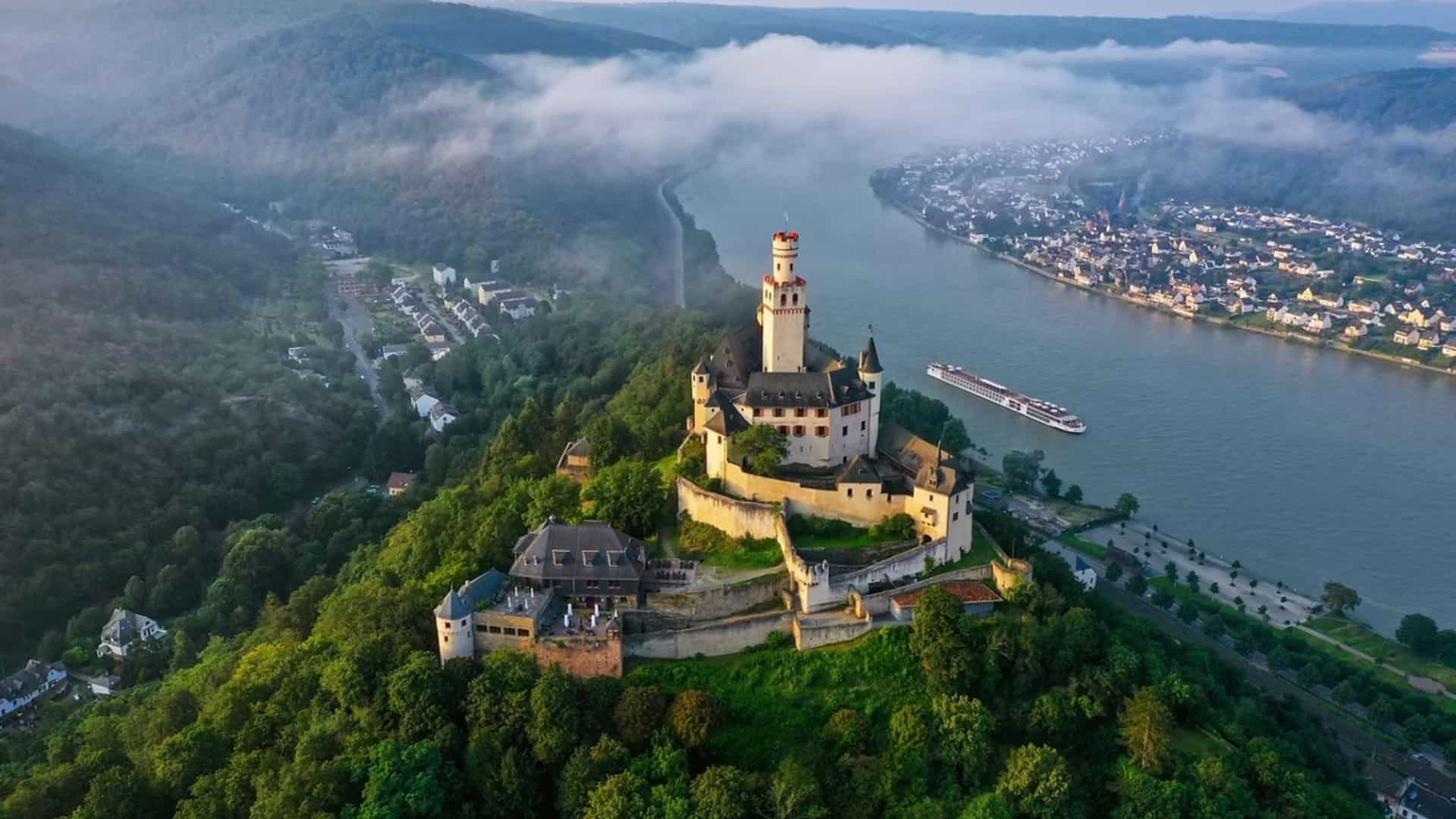 A Viking Longship sailing along the Rhine River past Marksburg Castle on a misty morning in Braubach, Germany, with the town and forested hillsides visible below.