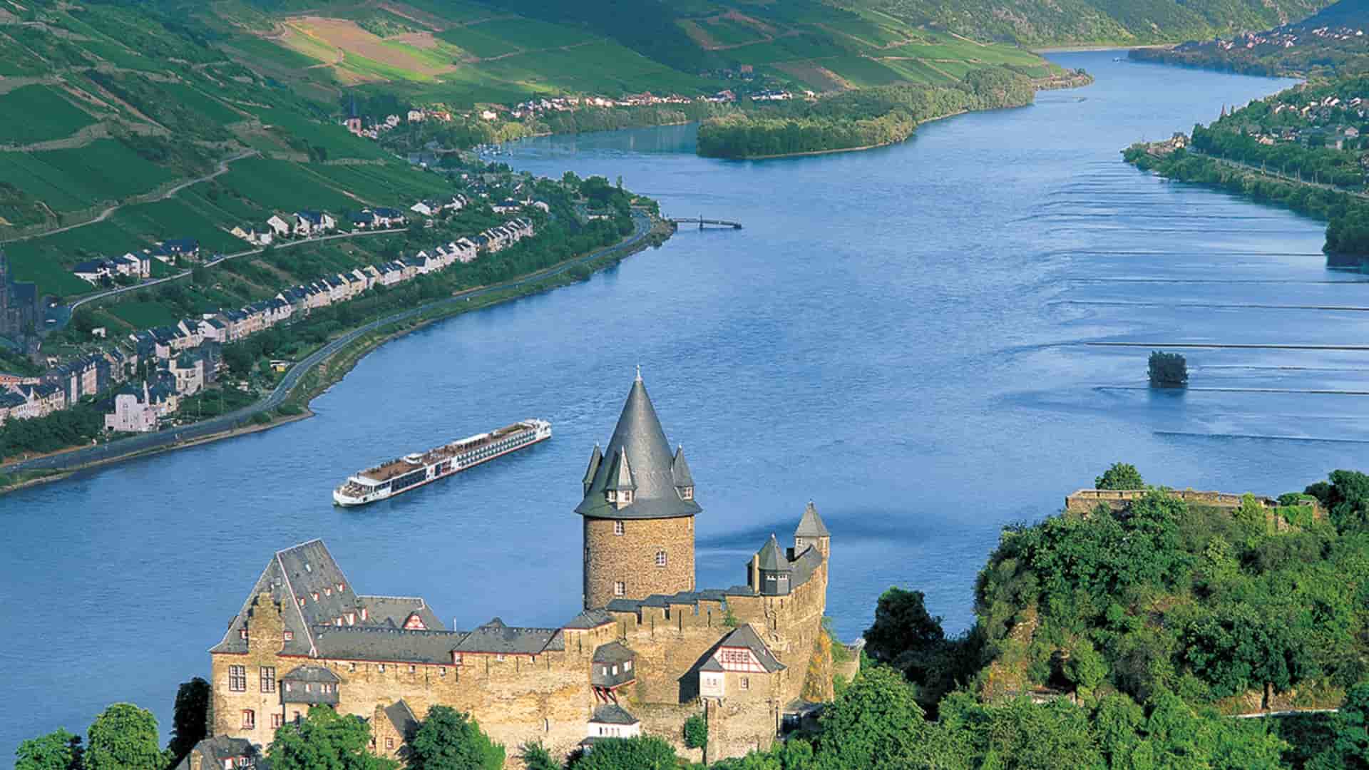 The historic Stahleck Castle overlooks a river cruise ship sailing along the winding Rhine River past lush, terraced vineyards in Bacharach, Germany.