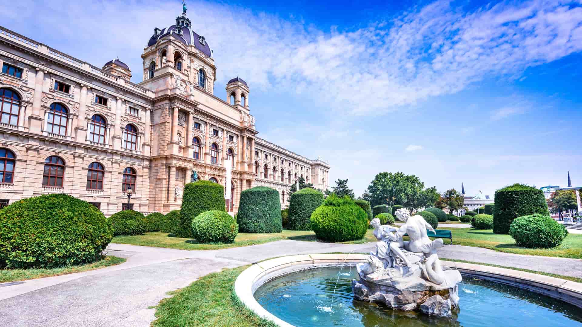 Grand historic architecture of a museum in Vienna, Austria, featuring a classical fountain with statues and neatly trimmed green bushes in the foreground under a bright blue sky.