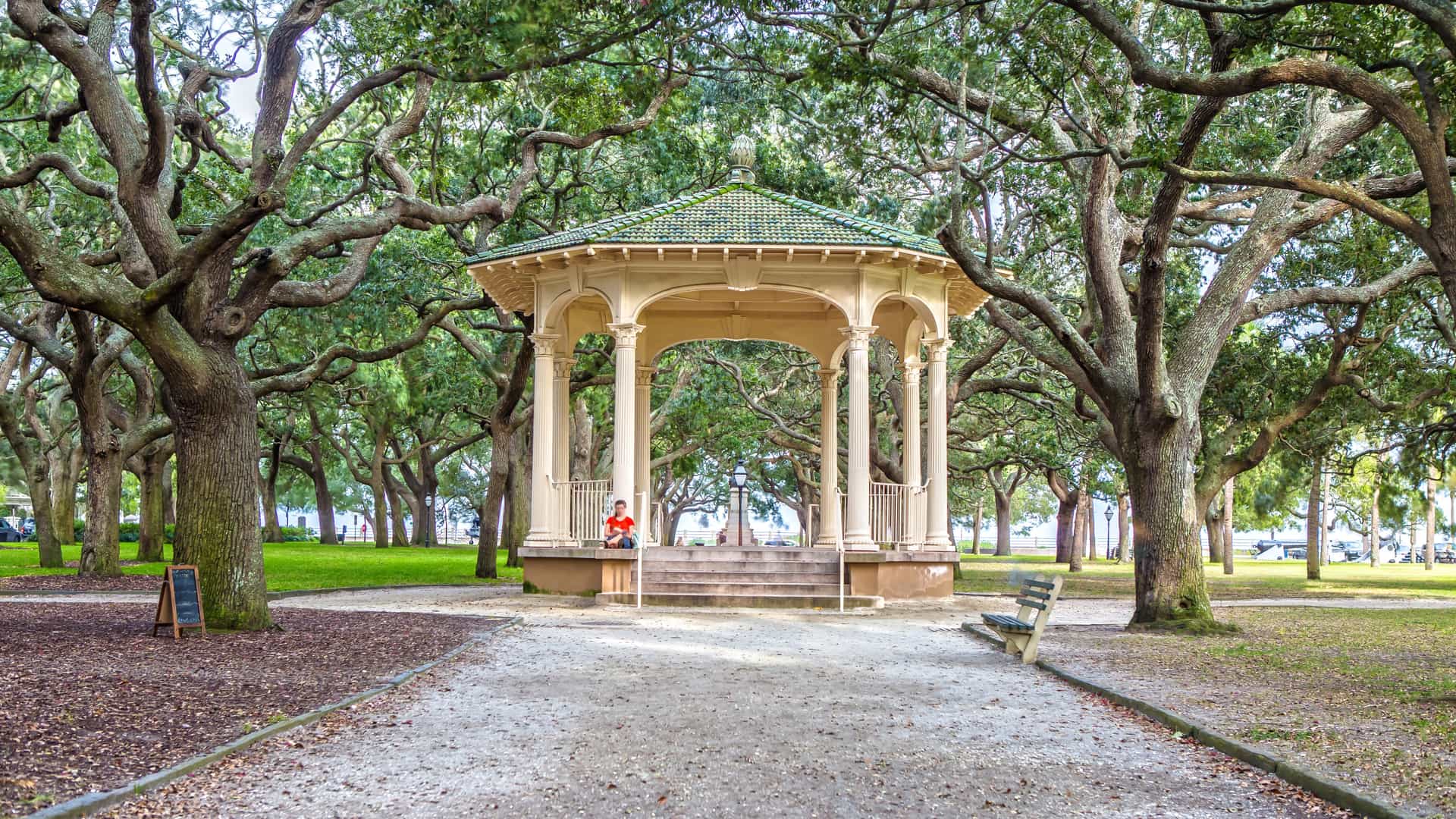 An elegant white gazebo stands centered under the sprawling canopy of ancient live oak trees in a sunlit, historic southern park.