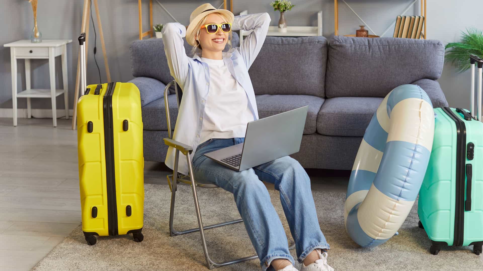 Woman sitting in a relaxed posed with her arms up and behind her head and with her laptop open, while there are suitcases packed on either side of her.